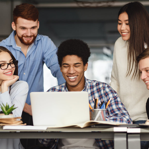 Group of five friends or students gathered around a laptop, smiling and engaged in a shared activity.