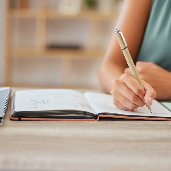 Person writing in a notebook with a pen on a wooden desk.