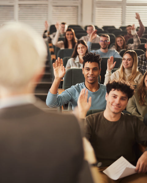 Classroom setting with students raising hands, facing a teacher.
