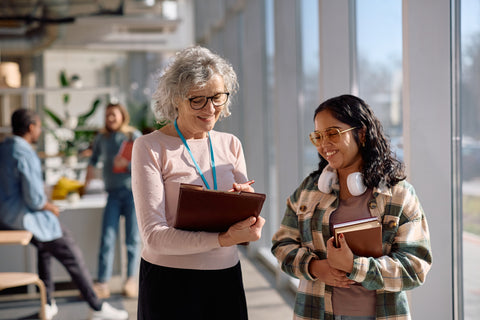 Two women in a modern office setting, one holding a folder and the other a notebook, with blurred background of people and plants.