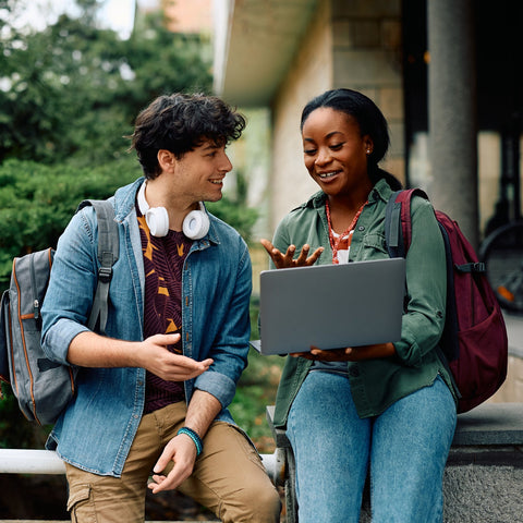 Two students sitting outdoors, one using a laptop, with a casual setting.