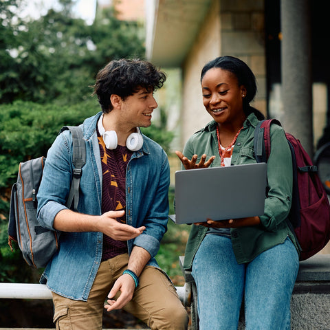 Two students sitting outdoors, one using a laptop, with a casual setting.