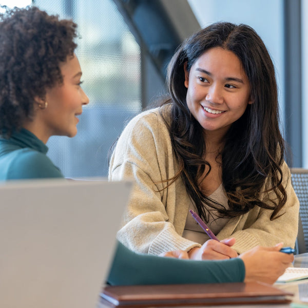 Two women sitting at a table with a laptop, engaged in a conversation.