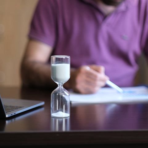 Person sitting at a desk with a laptop, hourglass, and pen, indicating a work or study session.