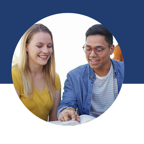 A young woman and a young man smiling as they look together at an open book, with him pointing to a passage on the page.