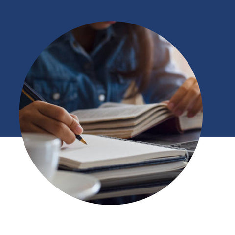 Close-up of a hand writing in a notebook beside open books and a cup of coffee.