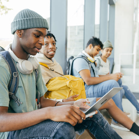 Diverse group of students sitting together indoors, using a tablet and mobile devices, engaged in digital learning and studying.