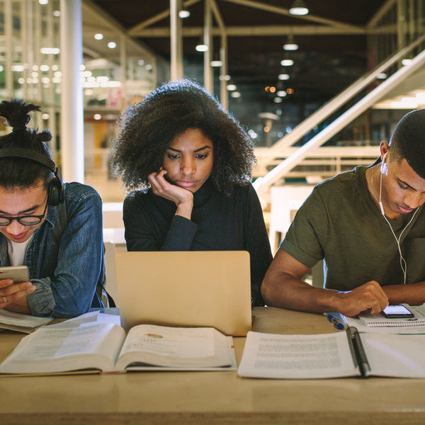 Multi-ethnic group of college students studying together at a table in a modern library, using laptops, books, and smartphones.