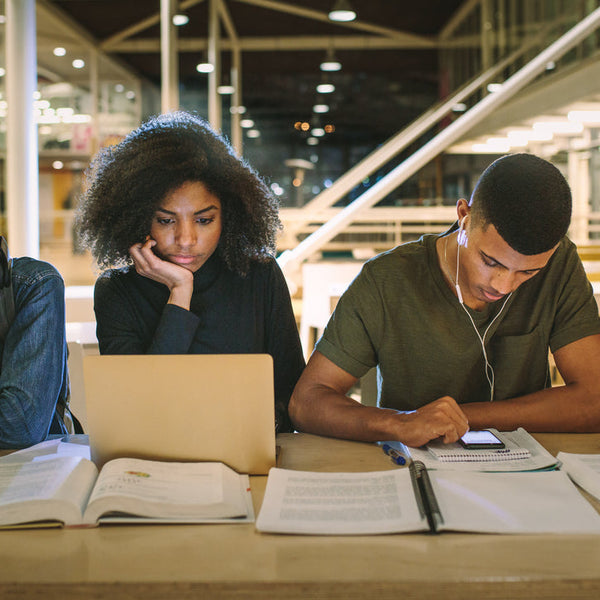 Multi-ethnic group of college students studying together at a table in a modern library, using laptops, books, and smartphones.
