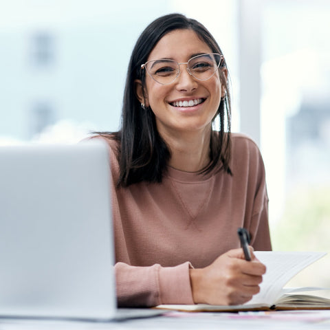 Smiling adult student studying at home with a laptop, writing in a notebook in a bright, comfortable workspace.