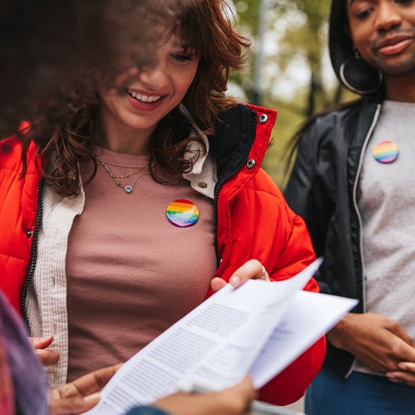 Two people outdoors, one wearing a red jacket with a rainbow flag, holding papers.