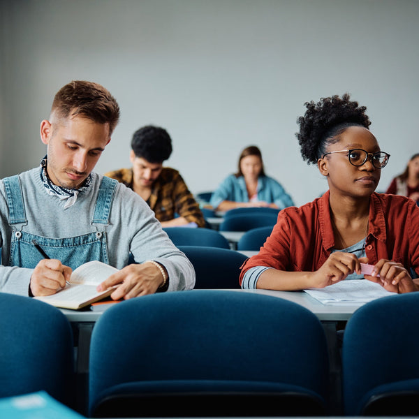 Multiracial group of college students seated in a classroom, taking notes and listening during a lecture, representing a campus learning environment and student engagement.