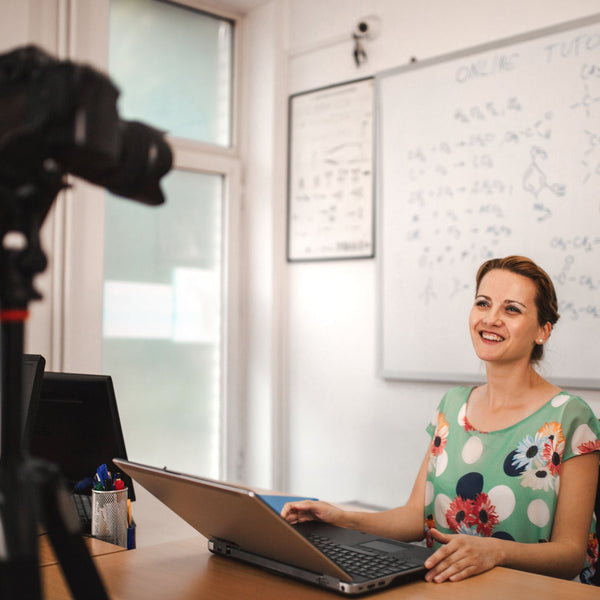 Woman sitting at a desk with a laptop, smiling, in front of a camera on a tripod. She is in an office setting with a whiteboard in the background.