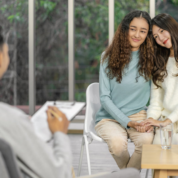 Two women sitting together in a room with a third person, possibly in a professional setting.