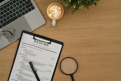 Resume on a clipboard with a laptop, coffee, and magnifying glass on a wooden desk.