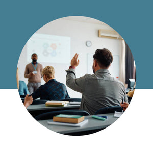 Group of people in a classroom setting with one person pointing at a whiteboard.