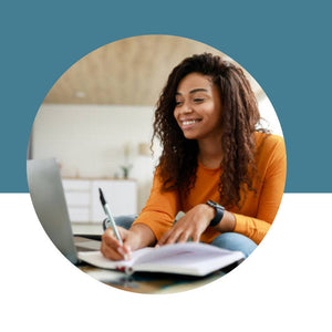 Smiling woman in an orange sweater writing in a notebook in front of a laptop on a teal and white background.