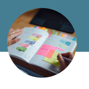 Hand writing in a planner filled with colorful sticky notes while another hand holds it open, on a teal and white background.