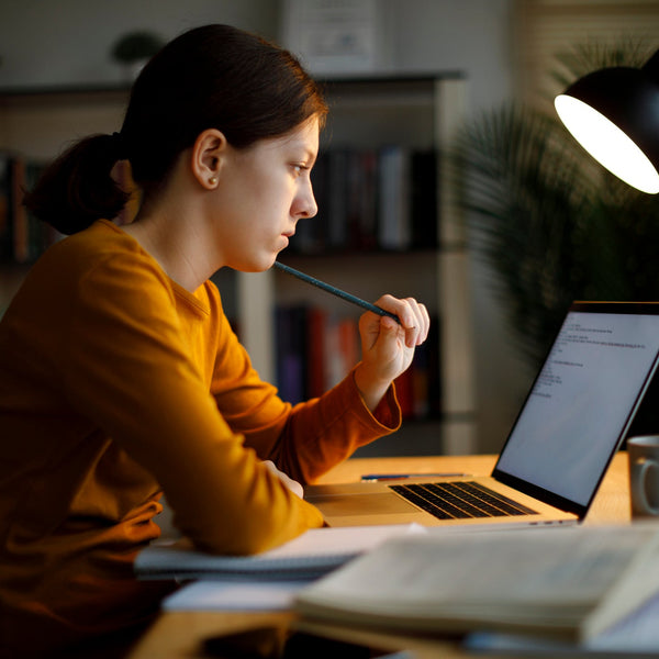 Student studying at a desk at night, thoughtfully reviewing content on a laptop while holding a pencil, focused and engaged.