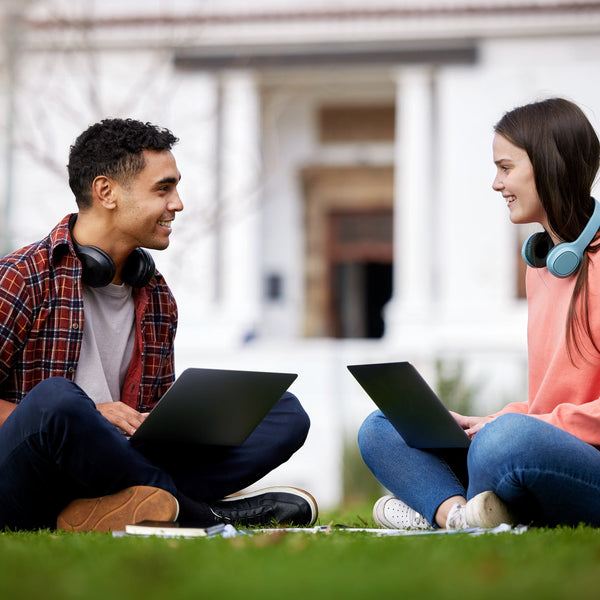 Two students sitting on grass with laptops in front of a building