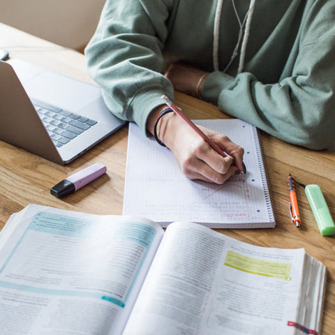Student studying at a desk with a laptop and open textbook, writing notes in a notebook, representing focused learning and effective study habits.