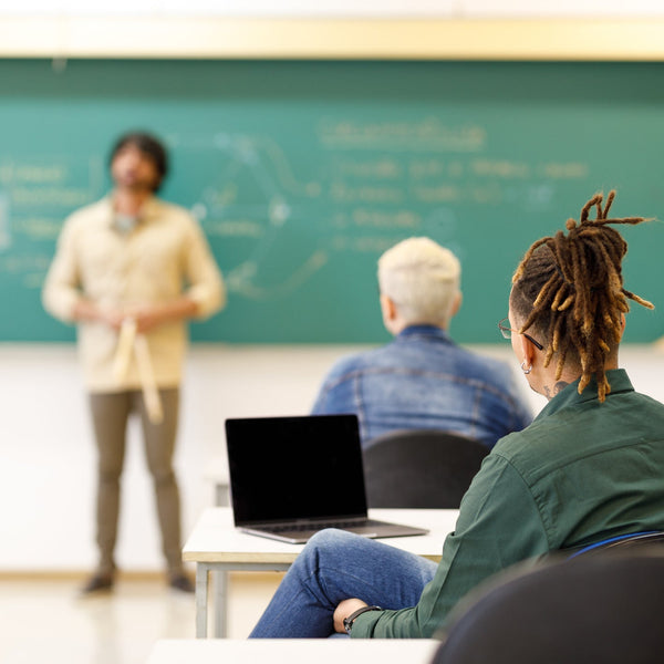 Person sitting at a desk with a laptop in front of a chalkboard in a classroom setting.