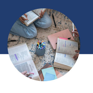 An overhead view of someone surrounded by open books, colorful pens, and notebooks while studying on the floor.
