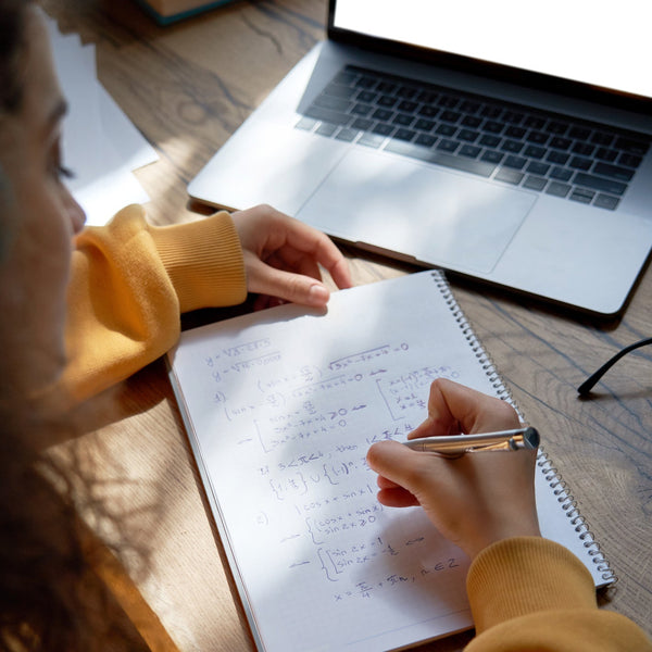 Student studying at a desk, writing notes in a notebook while using a laptop for online learning.