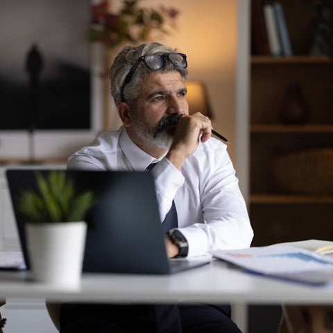 Thoughtful professional sitting at a desk with a laptop, looking away while reflecting, with a pen in hand in a home office setting.