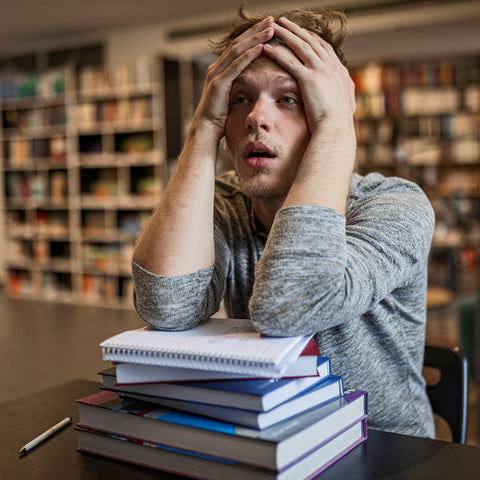 Overwhelmed college student sitting at a table with a stack of books, holding his head in a library setting.