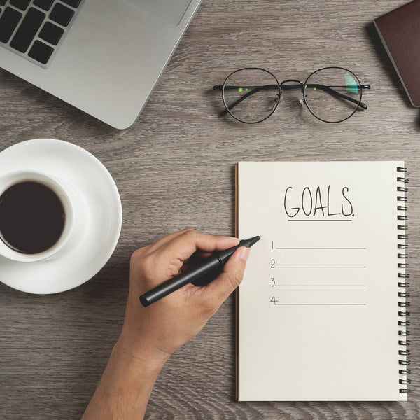 Person holding a pen over a notepad titled 'Goals' on a desk with a laptop, coffee, glasses, and a gift.