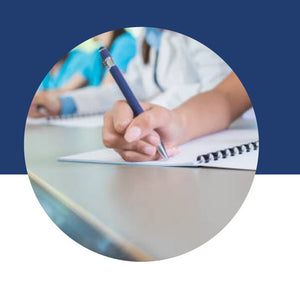 Close-up of a student's hand writing in a spiral notebook, with classmates working nearby.