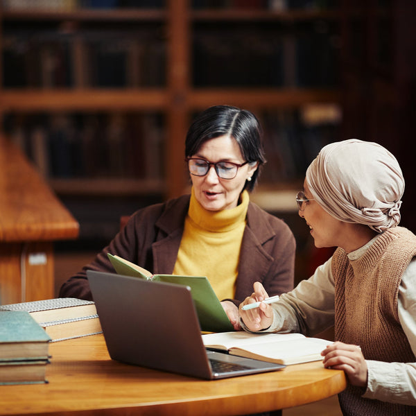 Two women sitting at a table with books and a laptop in a library setting.