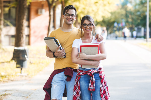 Male and female college students smiling and standing close, holding books on a campus path.

