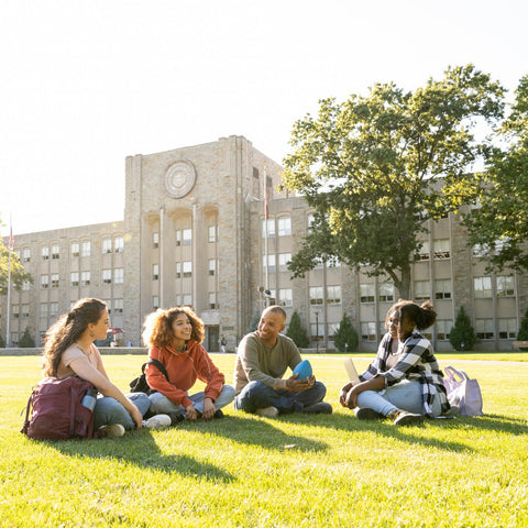 Four students sitting on a grassy area in front of a school building.
