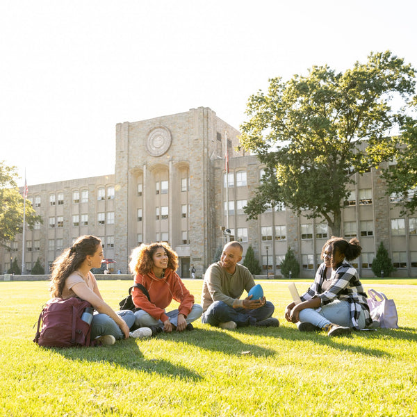 Four college students sitting together on a campus quad, talking and laughing in front of an academic building on a sunny day.