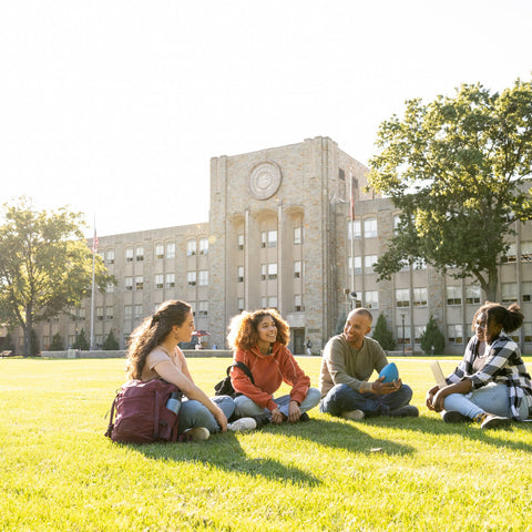 Four students sitting on a grassy area in front of a school building.
