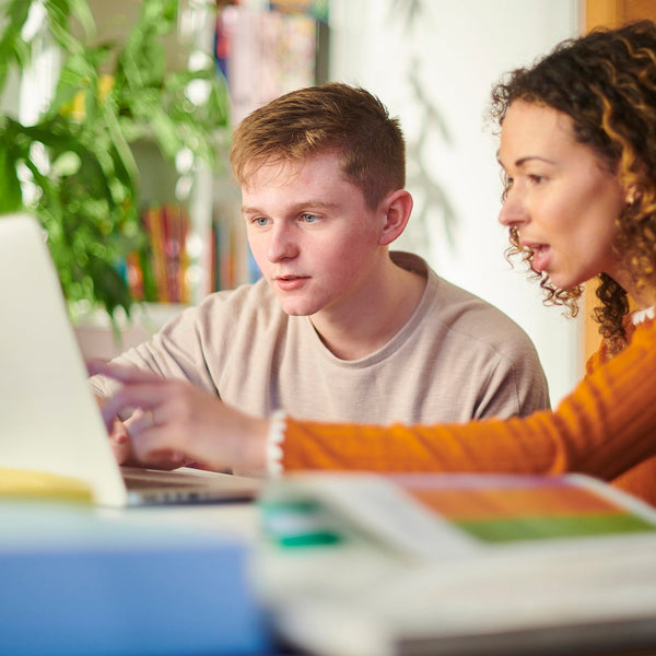 Two people working together on a laptop in a casual setting with books and plants in the background.