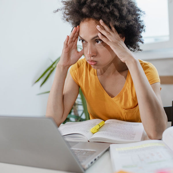 Woman in yellow shirt looking frustrated while using a laptop with books and stationery around.