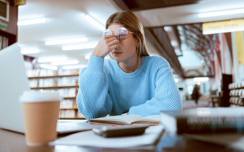 Woman in a library with books and a coffee cup, appearing stressed.