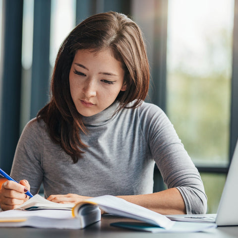 College student studying at a desk, writing notes in a notebook with a laptop nearby, focused and engaged in learning.