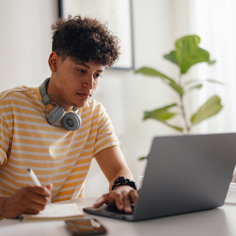 Male college student sitting at a desk with a laptop, writing in a notebook, and wearing headphones.