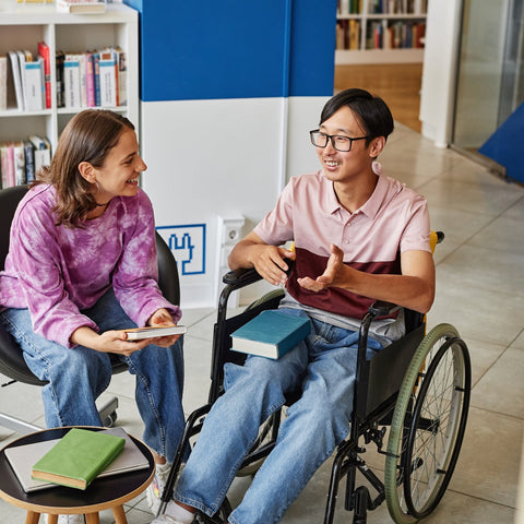Two college students talking and studying together in a library, including a student using a wheelchair, engaged in a friendly and collaborative discussion.