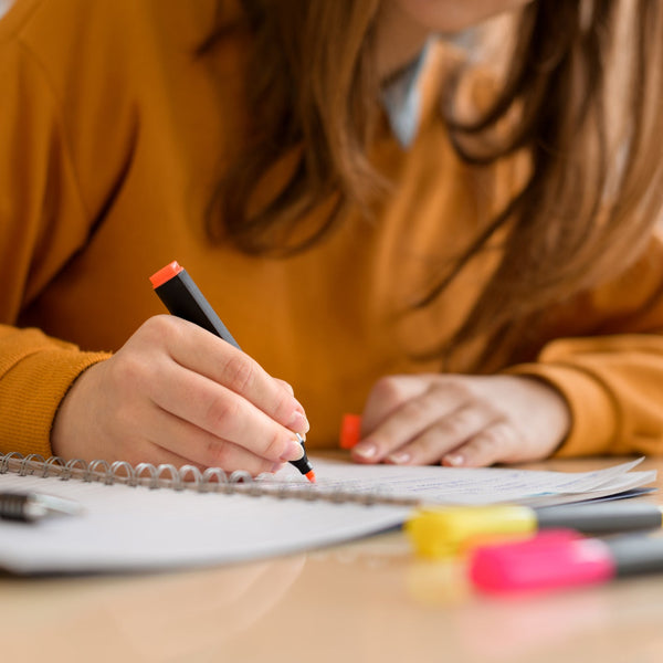 Person writing in a notebook with markers on a desk