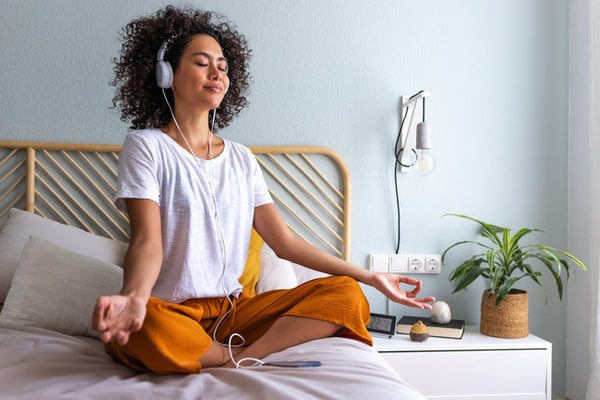 Woman meditating on a bed with headphones on, in a bedroom setting.