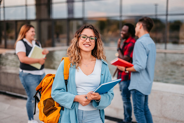 A young woman with glasses and a yellow backpack holds notebooks and smiles at the camera while three other people talk in the background outside a building.