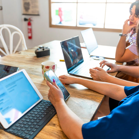People using digital devices including laptops, tablets, and smartphones at a table.