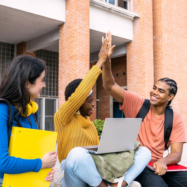 Three students sitting outside a brick building, with one using a laptop and another giving a high-five.