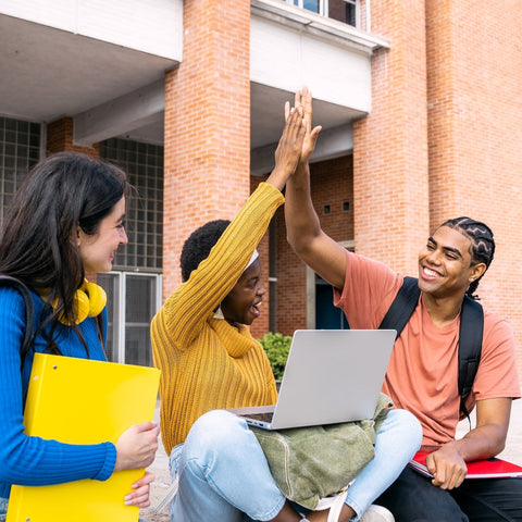 Three students sitting outside a brick building, with one using a laptop and another giving a high-five.