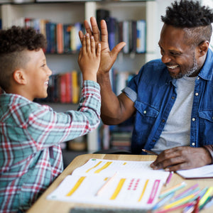 A father and son high-five over a desk with papers and colored pencils, set against a blurred background of a bookshelf.